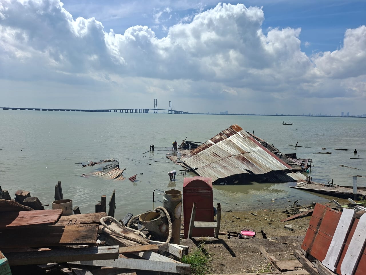 Dermaga Rindu di Kecamatan Labang, Bangkalan, tampak roboh dan tercebur ke laut setelah diterjang angin kencang dan hujan deras. Sejumlah warga terlihat memeriksa sisa bangunan yang rusak dengan latar Jembatan Suramadu di kejauhan. (doc. pribadi)